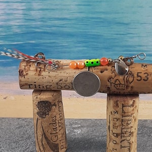 May include: A fishing lure with a red, orange, green and black bead, a silver blade and a feather tail. The lure is displayed on top of two wine corks.