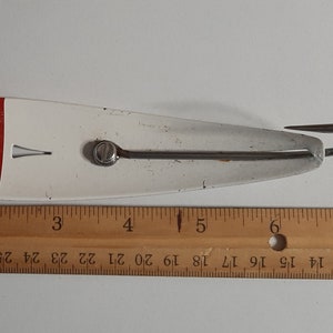 May include: A red and white fishing lure with a single hook. The lure is about 5 inches long and is measured against a ruler.