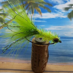 Puede incluir: Un señuelo de pesca con mosca verde y negro con una cola larga y plumosa. El señuelo está atado a un corcho con un alambre negro.