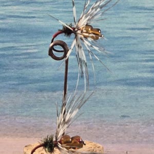 Könnte beinhalten: Zwei künstliche Angelköder. Eine Fliege ist auf einem Metallständer montiert, die andere ruht auf einem Korksockel. Beide Fliegen haben braune Körper, rote Akzente und weiß-schwarze Federflügel. Der Hintergrund ist ein verschwommenes Bild von Strand und Meer.