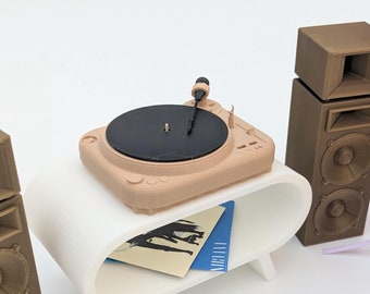 Può includere: Un giradischi in miniatura con due altoparlanti marroni, un supporto bianco e una pila di dischi in vinile. Il giradischi è su un supporto bianco con un bordo curvo. Gli altoparlanti sono rettangolari e hanno una finitura in legno marrone. I dischi in vinile sono impilati uno sopra l'altro e hanno diverse copertine di album. Il giradischi sta riproducendo un disco e l'ago si muove attraverso il disco.