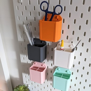 Puede incluir: Cinco organizadores de pared impresos en 3D en diferentes colores, incluyendo naranja, negro, blanco, rosa y azul claro. Los organizadores están montados en una pared de tablero de clavijas blanco y cada uno tiene varios compartimentos para guardar objetos pequeños.