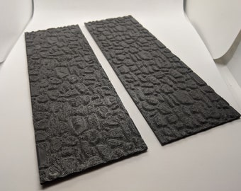 Puede incluir: Dos piezas rectangulares negras texturizadas que parecen piedra. El material está texturizado para parecer un camino de adoquines.