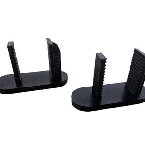 May include: Two black plastic phone stands with a textured surface. Each stand has two vertical supports with a grooved design and a flat base.
