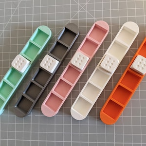 Puede incluir: Cinco bandejas de plástico de diferentes colores con cinco compartimentos cada una. Cada bandeja tiene un dado de plástico blanco con puntos negros en la parte superior.