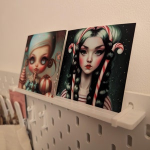 May include: Two illustrated prints of girls with candy cane hair. The girl on the left is wearing a blue and white dress and holding a candy cane ball. The girl on the right is wearing a red and white striped shirt and has a green background.