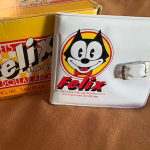 Könnte beinhalten: Eine weiße Felix-the-Cat-Geldbörse mit einem roten und gelben Felix-the-Cat-Logo. Die Geldbörse hat einen Druckknopfverschluss und den Text "1983 Felix The Cat Productions King Features Syndicate" unten aufgedruckt.