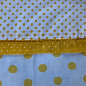 May include: Close-up of white fabric with a repeating pattern of yellow polka dots. A yellow band with small white dots separates the top and bottom sections. The bottom section features larger yellow polka dots on a white background. The fabric appears to be cotton.