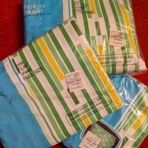 Op de afbeelding: Volledige lakensets in plastic verpakking. De lakens hebben een gestreept patroon in groen, geel en wit. De verpakking is blauw en wit, met de tekst "No Iron Muslin" en "Bright Mint".