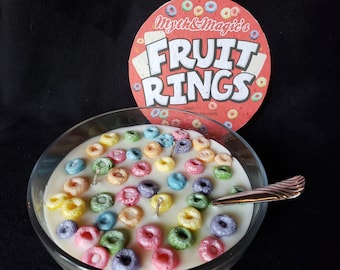 May include: A white candle shaped like a bowl of cereal with colorful fruit rings on top. The candle is labeled "Myth & Magic's Fruit Rings - Sweet & Fruity Candle".