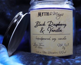 May include: A hand-poured soy candle in a glass jar with a black lid. The label is cream-colored with black text that reads "MYTH & Magic", "Black Raspberry & Vanilla", "handpoured soy candle", "Burn Time: 30 hours", "Net Wt. 7oz", and "made in New Hampshire, USA".