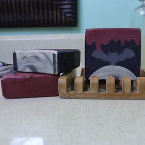 May include: Three bars of handmade soap, two are red and one is black with a white swirl design. The black soap has a bat silhouette on it. The soaps are on a bamboo soap dish.