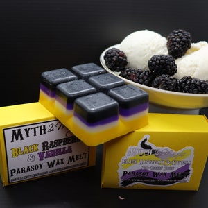 May include: Two yellow boxes with black text that reads "MYTH & MAGIC" and "BLACK RASPBERRY & VANILLA PARASOY WAX MELT". The boxes contain black and purple wax melts in a square shape. The wax melts are arranged in a grid pattern. The boxes are sitting on a white surface.