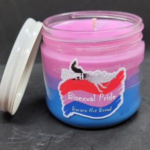 May include: A glass jar candle with a pink, purple, and blue layered wax design. The label reads "Myth & Magic" and "Bisexual Pride" with the scent "Banana Nut Bread".