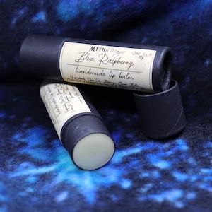 May include: Two black tubes of lip balm with white labels. The label on the top tube reads "Myth & Magic Blue Raspberry handmade lip balm".