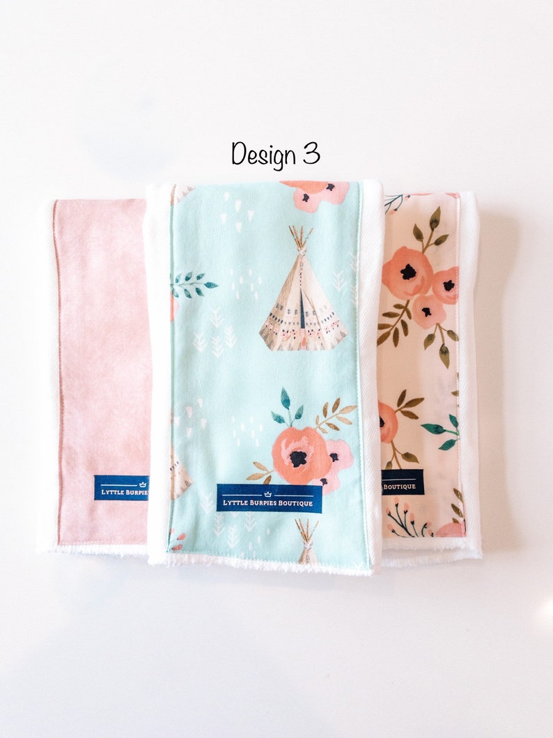 May include: Three burp cloths with different designs. The first is a solid pink, the second is a light blue with a teepee and floral pattern, and the third is a peach with a floral pattern. All three cloths have white trim and a blue label that says "LYTTLE BURPIES BOUTIQUE".
