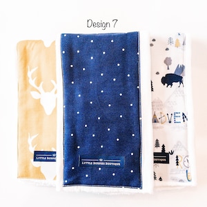 May include: Three baby burp cloths with different designs. The first cloth is yellow with a white deer silhouette. The second cloth is blue with white polka dots. The third cloth is white with a gray mountain scene and the word "Adventure". All three cloths have a white border and the text "Lyttle Burpies Boutique" on the bottom.