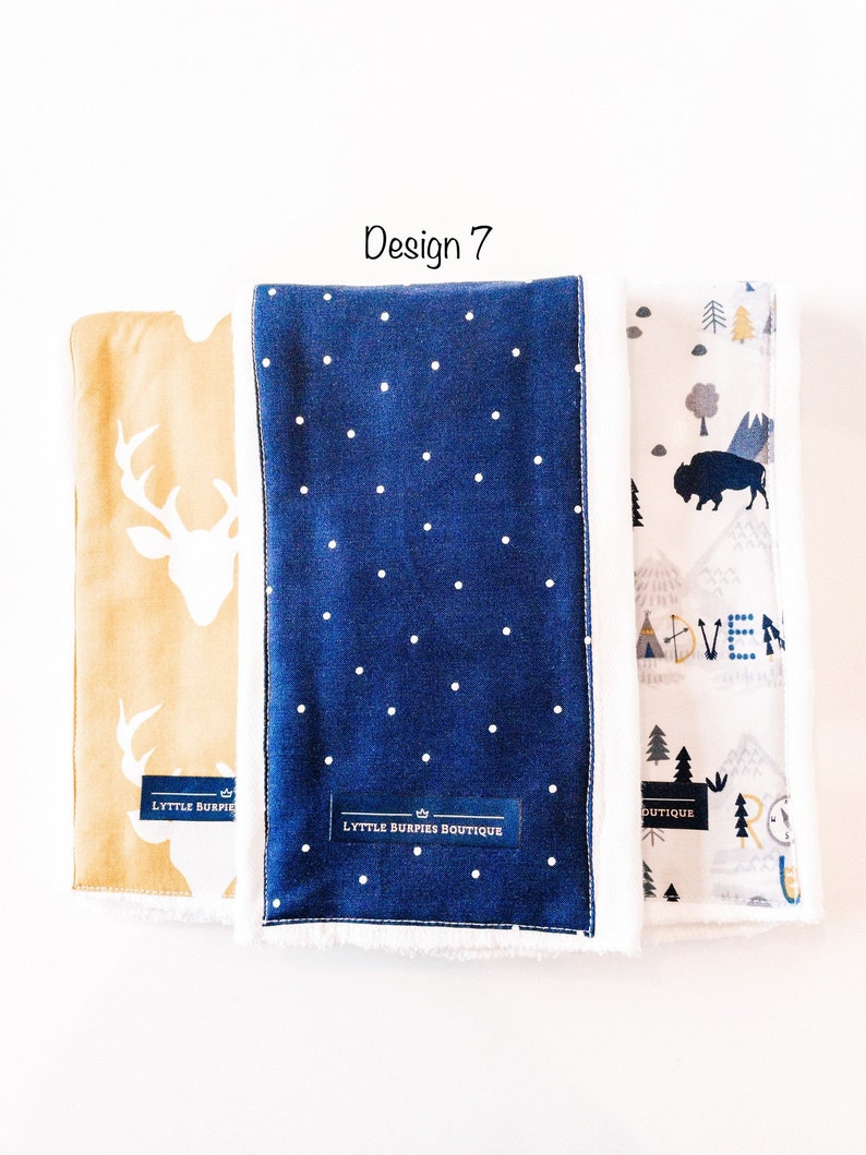 May include: Three baby burp cloths with different designs. The first cloth is yellow with a white deer silhouette. The second cloth is blue with white polka dots. The third cloth is white with a gray mountain scene and the word "Adventure". All three cloths have a white border and the text "Lyttle Burpies Boutique" on the bottom.