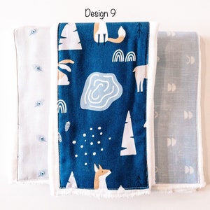 May include: Three fabric burp cloths with different patterns. The center cloth is blue with white trees, a fox, a deer, and a pond. The cloth on the left is white with blue dots. The cloth on the right is blue with white crescent moons.