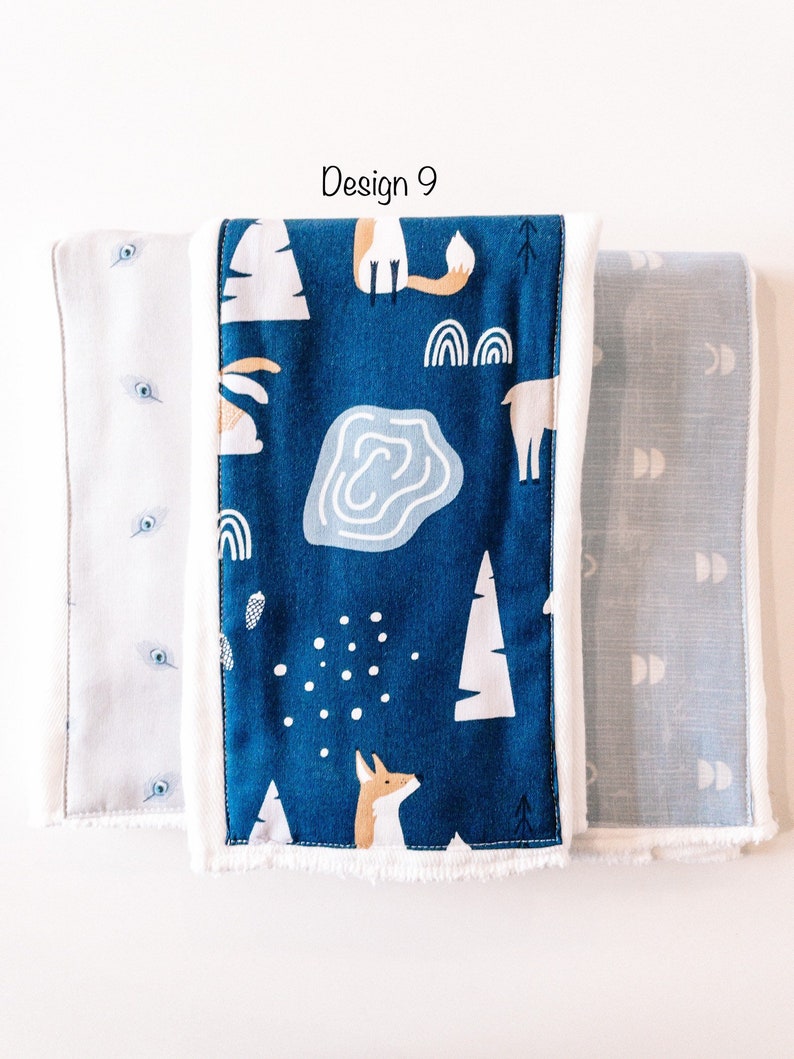 May include: Three fabric burp cloths with different patterns. The center cloth is blue with white trees, a fox, a deer, and a pond. The cloth on the left is white with blue dots. The cloth on the right is blue with white crescent moons.