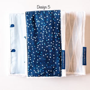 May include: Three burp cloths with different patterns. The first is light blue with a whale design. The second is dark blue with white polka dots. The third is light gray with a wavy pattern. All three cloths have white trim and the text "LITTLE BURPIES BOUTIQUE" is printed on the bottom right corner of each cloth.