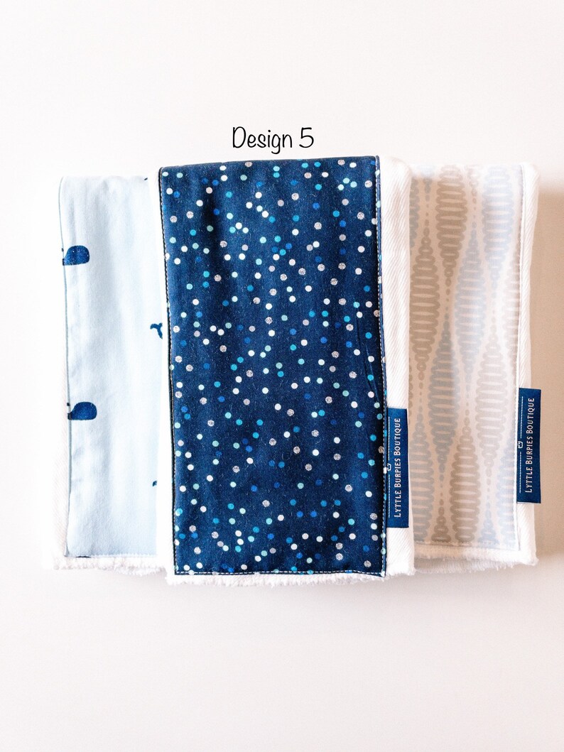 May include: Three burp cloths with different patterns. The first is light blue with a whale design. The second is dark blue with white polka dots. The third is light gray with a wavy pattern. All three cloths have white trim and the text "LITTLE BURPIES BOUTIQUE" is printed on the bottom right corner of each cloth.