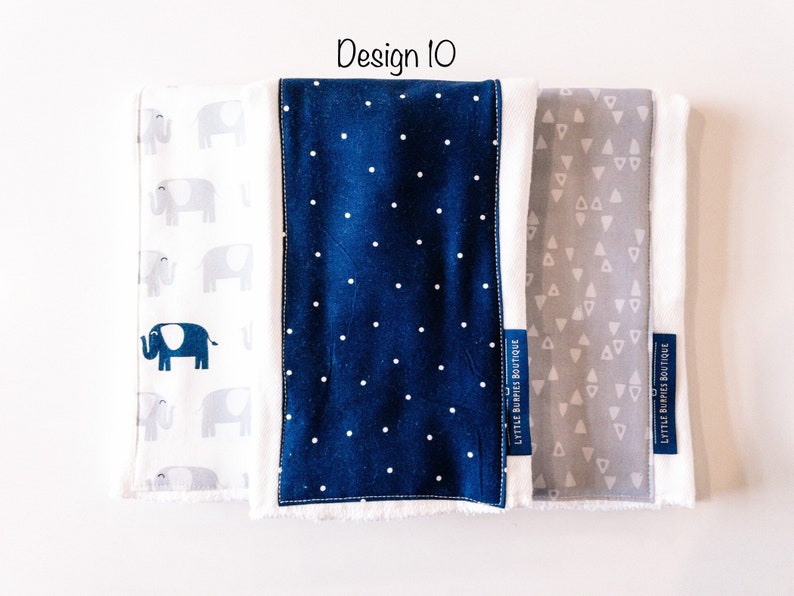 May include: Three fabric burp cloths with different patterns. The first cloth has a white background with gray elephants. The second cloth has a navy blue background with white polka dots. The third cloth has a gray background with white triangles. Each cloth has a white label with the text "Little Burpies Boutique".