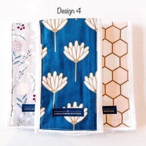 May include: Three burp cloths with different patterns. The first is a light gray with a floral design, the second is a teal blue with a white flower design, and the third is a peach with a honeycomb pattern. Each cloth has a white border and a label that says "Lyttle Burpies Boutique".