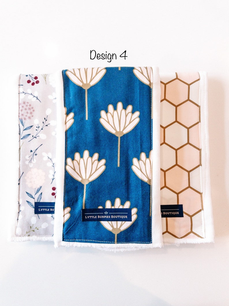 May include: Three burp cloths with different patterns. The first is a light gray with a floral design, the second is a teal blue with a white flower design, and the third is a peach with a honeycomb pattern. Each cloth has a white border and a label that says "Lyttle Burpies Boutique".