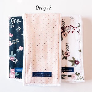 May include: Three burp cloths with different floral patterns. The cloths are white with a soft, white fleece backing. The center cloth has a pink background with small white and red dots. The cloth on the left has a dark blue background with white and pink floral designs. The cloth on the right has a white background with red, pink, and green floral designs. Each cloth has a blue label with the text "LYTTLE BURPIES BOUTIQUE" and a crown.