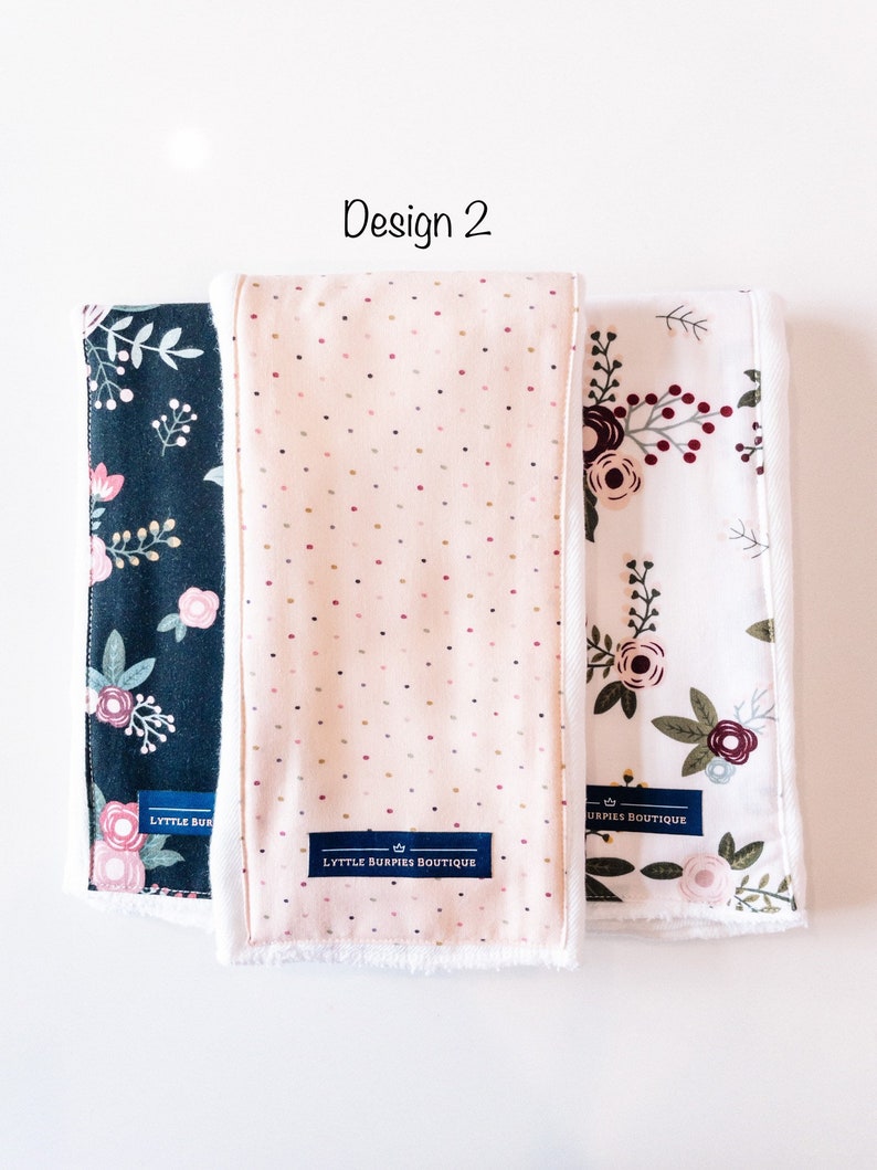 May include: Three burp cloths with different floral patterns. The cloths are white with a soft, white fleece backing. The center cloth has a pink background with small white and red dots. The cloth on the left has a dark blue background with white and pink floral designs. The cloth on the right has a white background with red, pink, and green floral designs. Each cloth has a blue label with the text "LYTTLE BURPIES BOUTIQUE" and a crown.