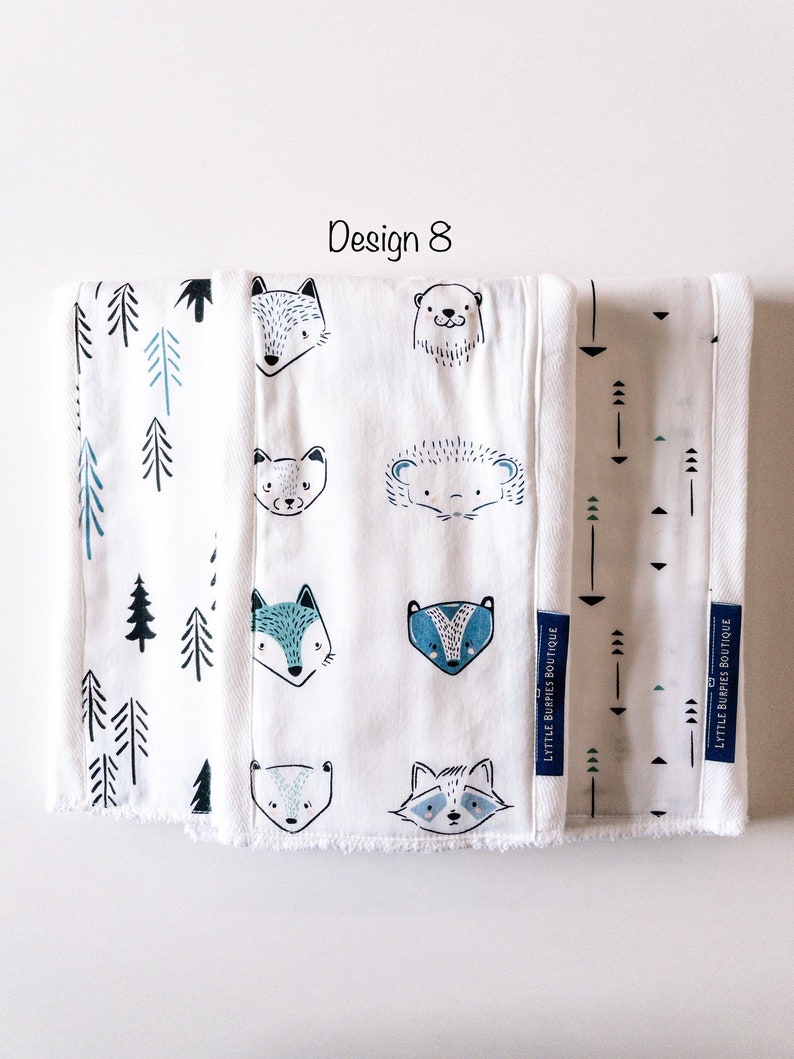 May include: Three white burp cloths with different designs. The first cloth has a blue and white pattern of trees. The second cloth has a blue and white pattern of animals, including a fox, a bear, a raccoon, a badger, and a hedgehog. The third cloth has a blue and white pattern of arrows. The text "LYTTLE BURPIES BOUTIQUE" is printed on the bottom right corner of each cloth.
