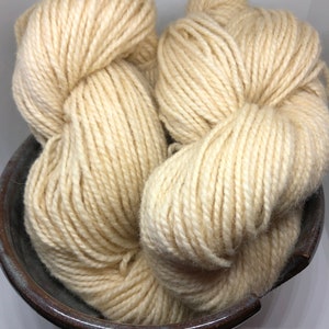 May include: Two skeins of off-white yarn are nestled in a dark brown ceramic bowl. The yarn appears thick and textured, with visible strands. The bowl has a glossy finish. This image is suitable for craft and knitting projects.
