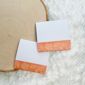 May include: Two white sticky notes with a border of orange and white fall leaves. The notes are on a white fuzzy surface.