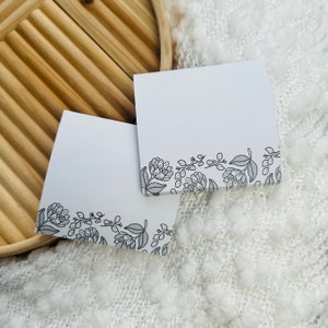 May include: Two white sticky notes with a black floral border design. The notes are on a white fuzzy surface.