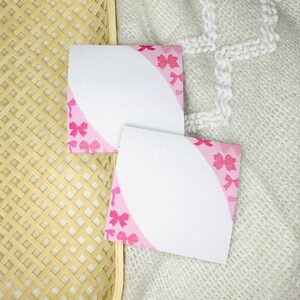 May include: Two blank white note cards with pink bows printed on the edges. The cards are on a woven wicker surface and a white knitted fabric.