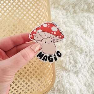 May include: A whimsical sticker featuring a cartoon mushroom with a red cap and white spots. The mushroom has a smiling face and the words "MAKE SOME MAGIC" in black lettering. The sticker is held against a woven background.