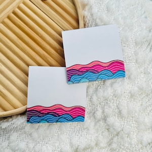 May include: Two white sticky note pads with a colorful abstract design of pink, purple, and blue waves at the bottom of each pad.