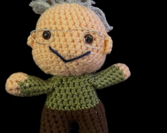 May include: A handmade crochet doll depicting an elderly person. The doll has a peach-colored head with gray hair, wire-rimmed glasses, and a stitched smile. It wears a green sweater and brown pants.