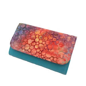 Könnte beinhalten: Eine türkisfarbene Brieftasche mit einer bunten, gemusterten Klappe. Die Klappe hat ein rotes, orangefarbenes, gelbes und violettes Muster aus Kreisen. Der Name "Yanna" ist in Rot auf die Klappe gestickt.