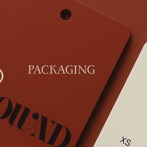 May include: Close-up of two rectangular tags. One is a deep red-brown color with the word "PACKAGING" in white text. The other tag is off-white with the word "CROWN" printed in black. The tags have a hole at the top.