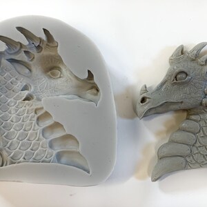 May include: A silicone mold and a gray resin cast of a dragon head. The dragon has a detailed face with scales and spikes.