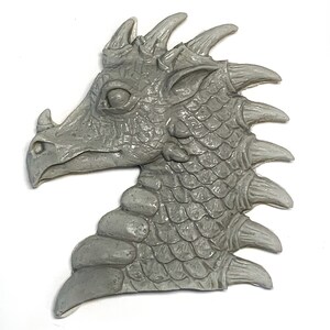 May include: A gray resin dragon head sculpture with detailed scales and spikes. The dragon's head is facing to the side, with its mouth closed and its eyes closed. The sculpture is a great addition to any fantasy or dragon-themed decor.