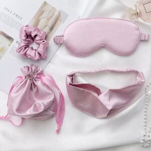 May include: A set of three pink satin accessories: a sleep mask, a headband, and a small drawstring bag. The items are arranged on a white surface.