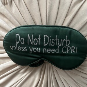 Peut inclure: Un masque de sommeil vert foncé avec le texte "Do Not Disturb unless you need CPR!" en paillettes argentées. Le masque a un élastique et repose sur une surface beige texturée. Le masque de sommeil est conçu pour bloquer la lumière.