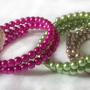 Op de afbeelding: Vier parel armbanden in verschillende kleuren: crème, roze, groen en bruin. Elke armband is gemaakt van twee rijen parels.