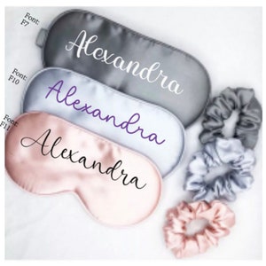 May include: Three satin sleep masks with the name "Alexandra" embroidered on them in different fonts and colours. The masks are in grey, light grey, and pink. There are also three matching scrunchies in grey and pink.