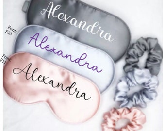 May include: Three satin sleep masks with the name "Alexandra" embroidered on them in different fonts and colors. The masks are in gray, light gray, and pink. There are also three matching scrunchies in gray and pink.