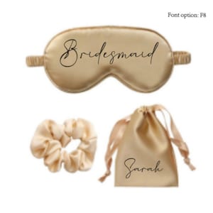 May include: A gold satin sleep mask with the word "Bridemaid" printed in black script, a gold satin scrunchie, and a gold satin drawstring bag with the name "Sarah" printed in black script.