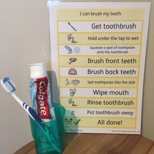 May include: A green plastic cup containing a toothbrush and a tube of Colgate toothpaste. A yellow and white poster with the text "I can brush my teeth" and a list of steps for brushing teeth.